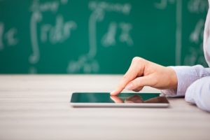 Flexible Teaching in 2017 teachers close up of teacher's hand touching a tablet on a desk in a classroom with a blackboard and writing in the background