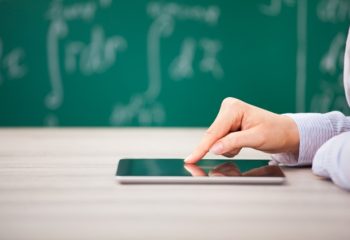 Flexible Teaching in 2017 teachers close up of teacher's hand touching a tablet on a desk in a classroom with a blackboard and writing in the background